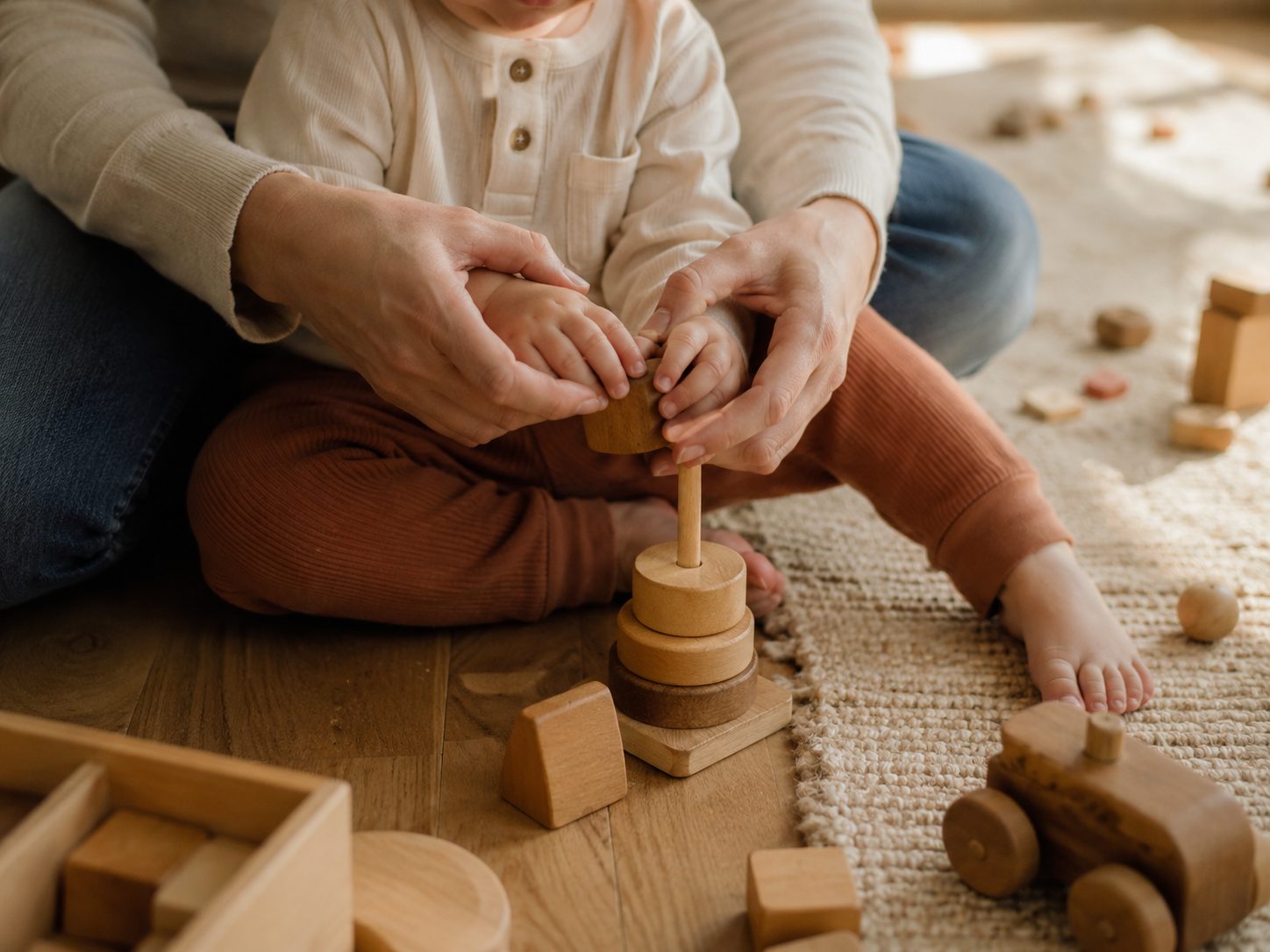 A parent&rsquo;s hands guiding a toddler stacking wooden toys on the floor